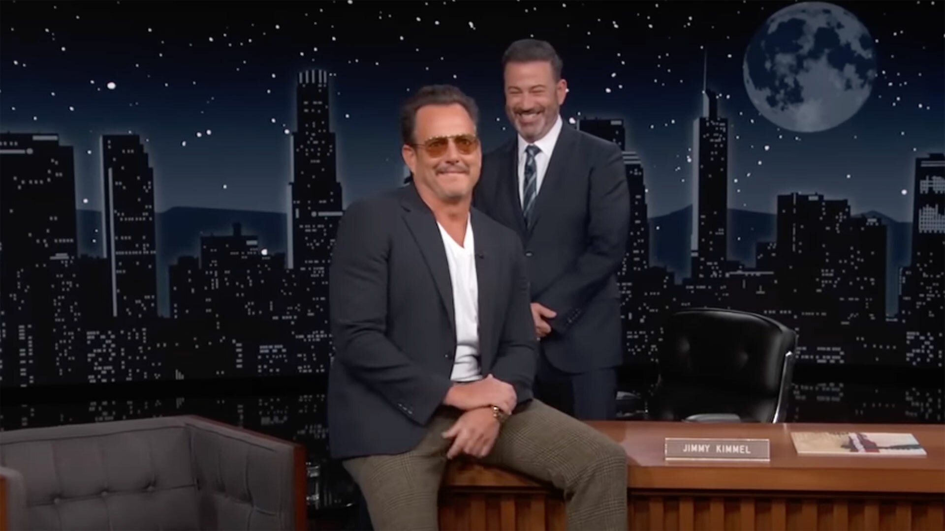 A man in sunglasses sits on a talk show desk while another man in a suit stands behind laughing.