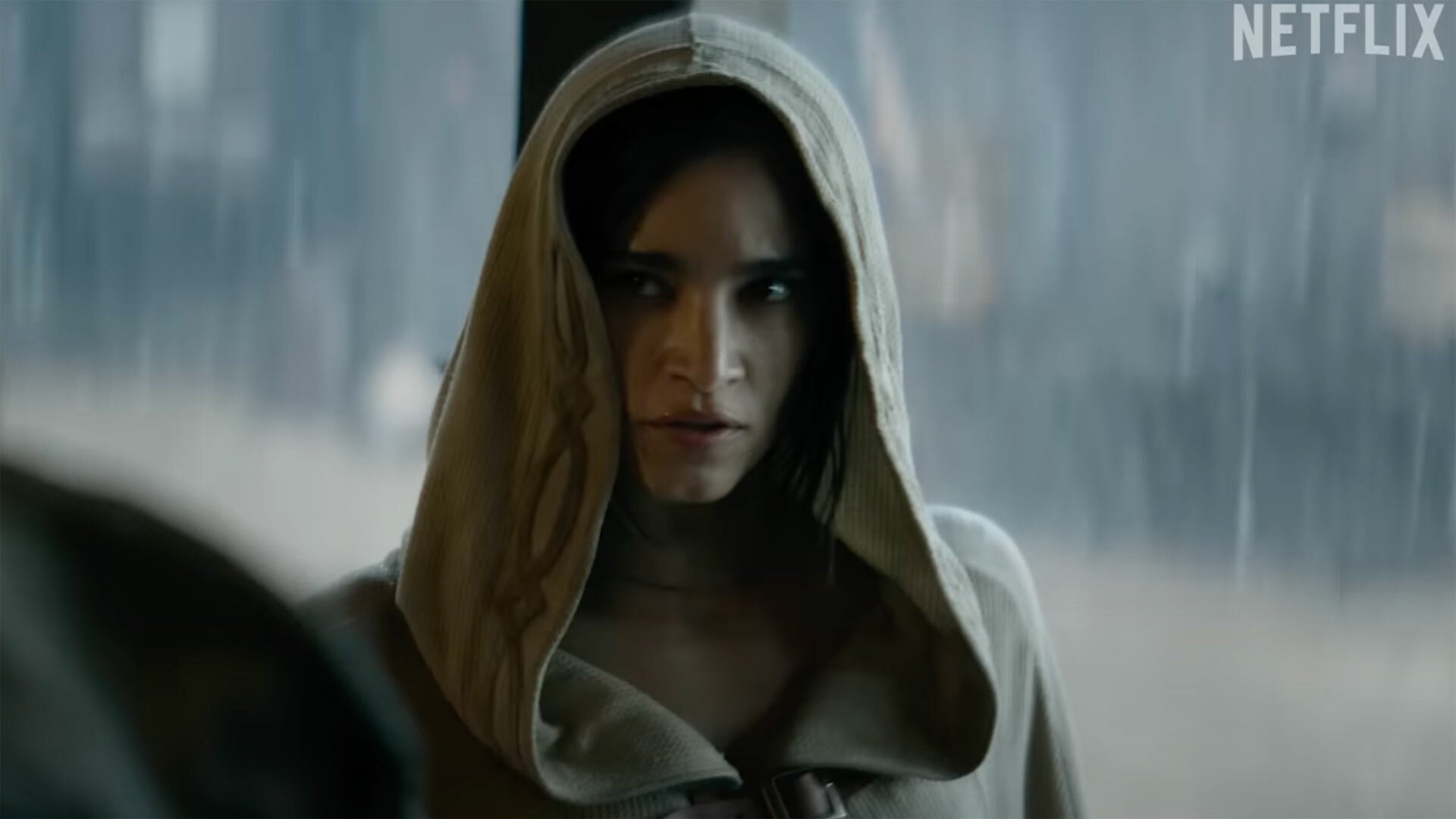 A woman in a grey cloak with the hood up stands indoors, while rain pounds down outside.