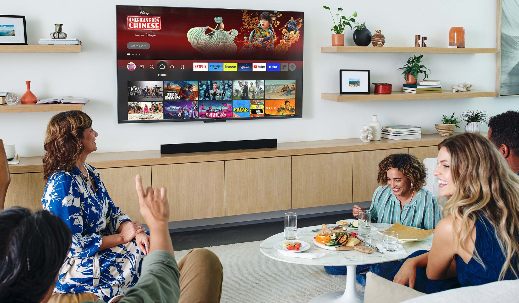 Four people sitting in living room with snacks and TV on wall with streaming apps on screen