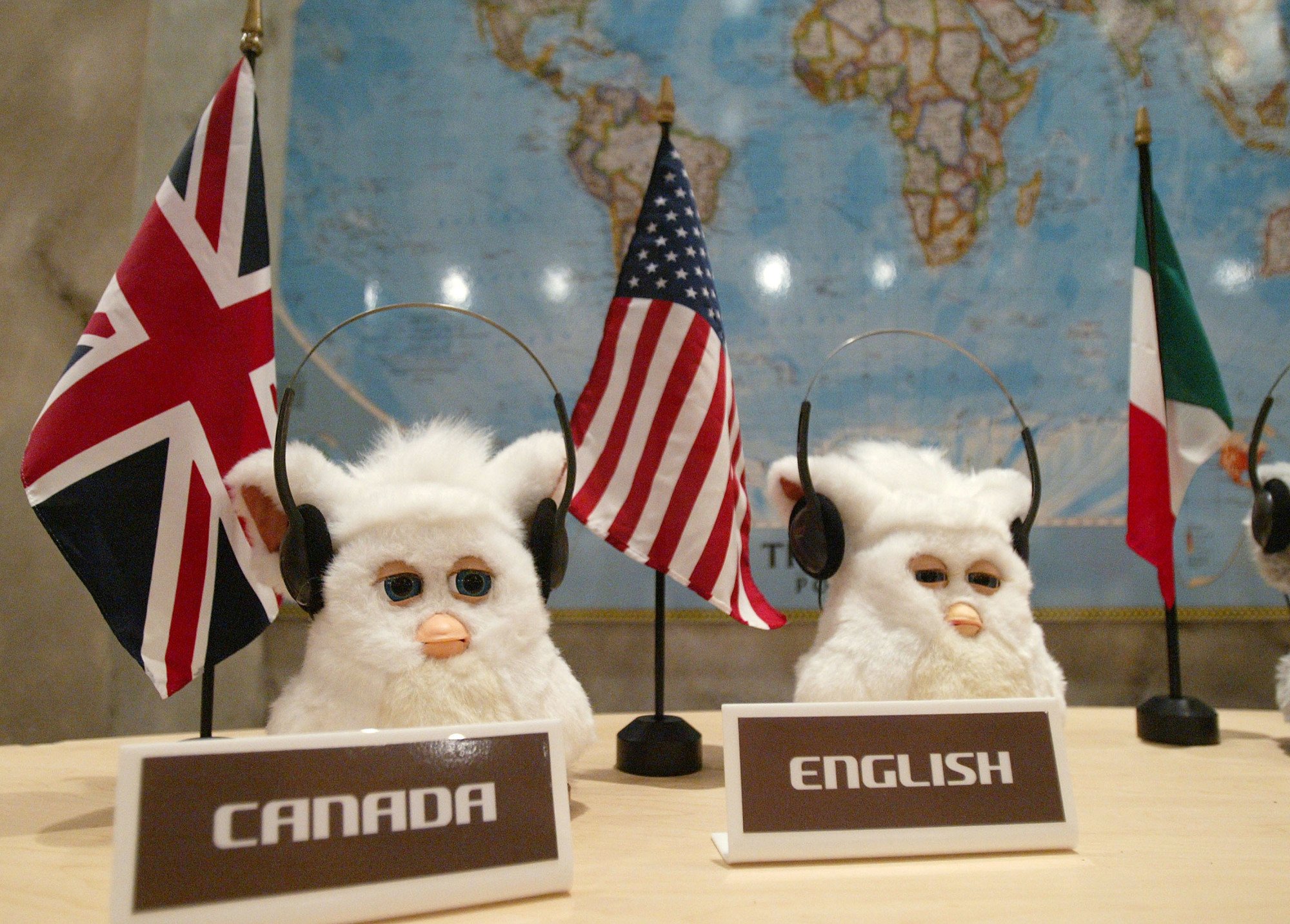 Two Furbies sitting at a desk, wearing headphones. Behind them are several flags from different countries, and in front of them are title plaques labelled "Canada" and "English."