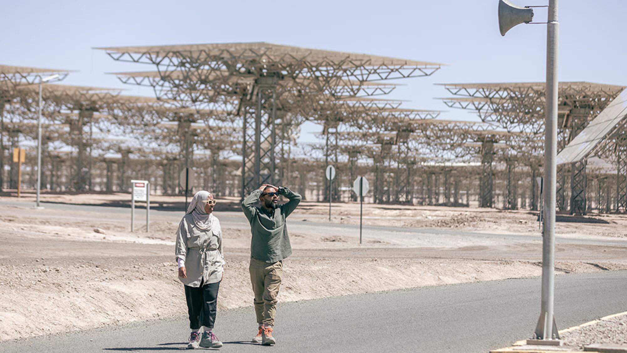 A man and woman walk through the desert with metal structures in the background.
