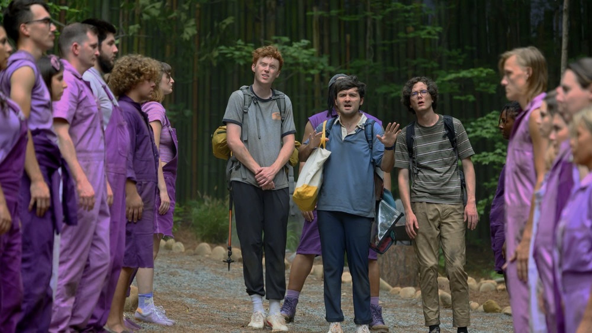 Three young men in hiking clothes walk through a crowd of people in purple clothes.