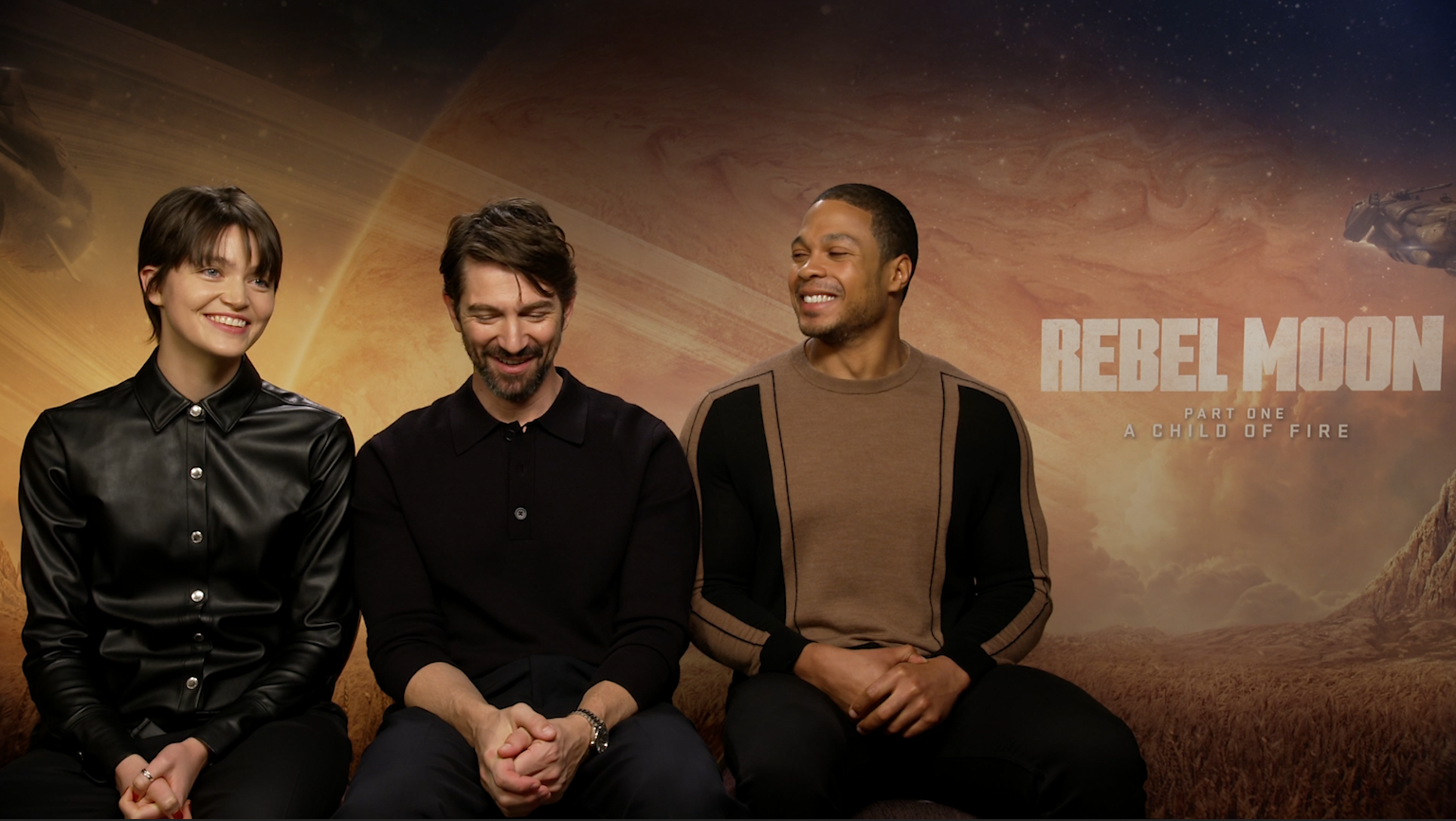 'Rebel Moon' actors E. Duffy, Michiel Huisman, and Ray Fisher (left to right) are sit against a Rebel Moon promo backdrop. All three are laughing and looking at each other