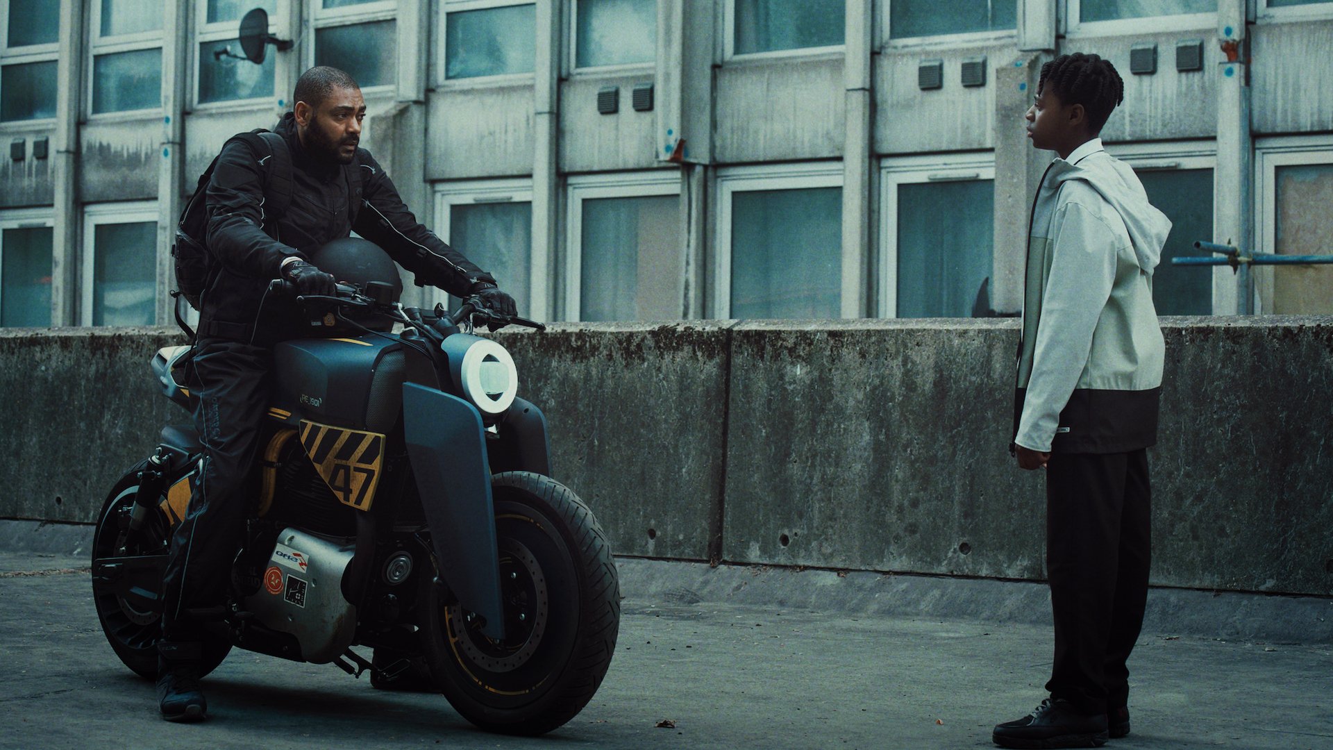 A man on a motorbike faces a young boy on a grey-lit street.