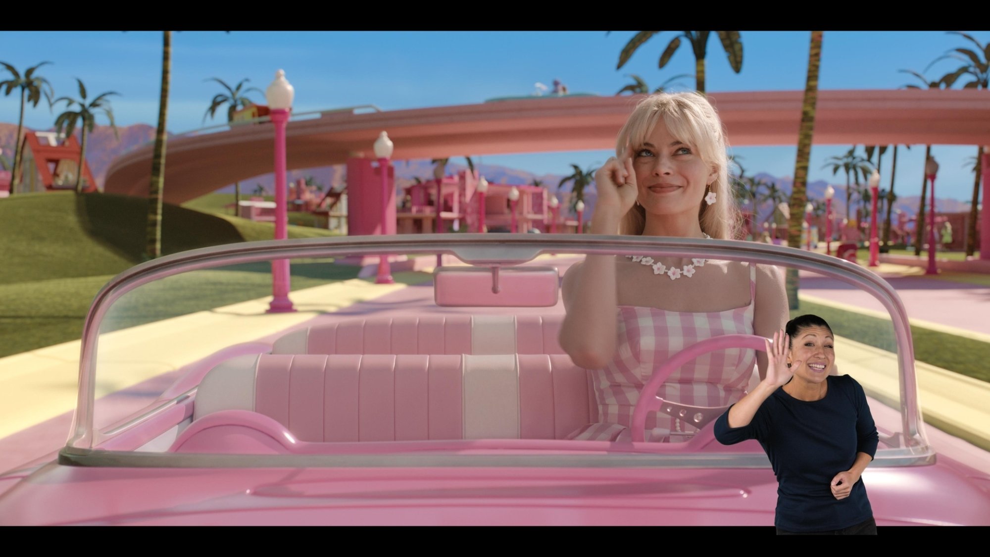 Margot Robbie as Barbie drives a pink convertible. A woman in a black shirt interprets in ASL in the bottom right corner.