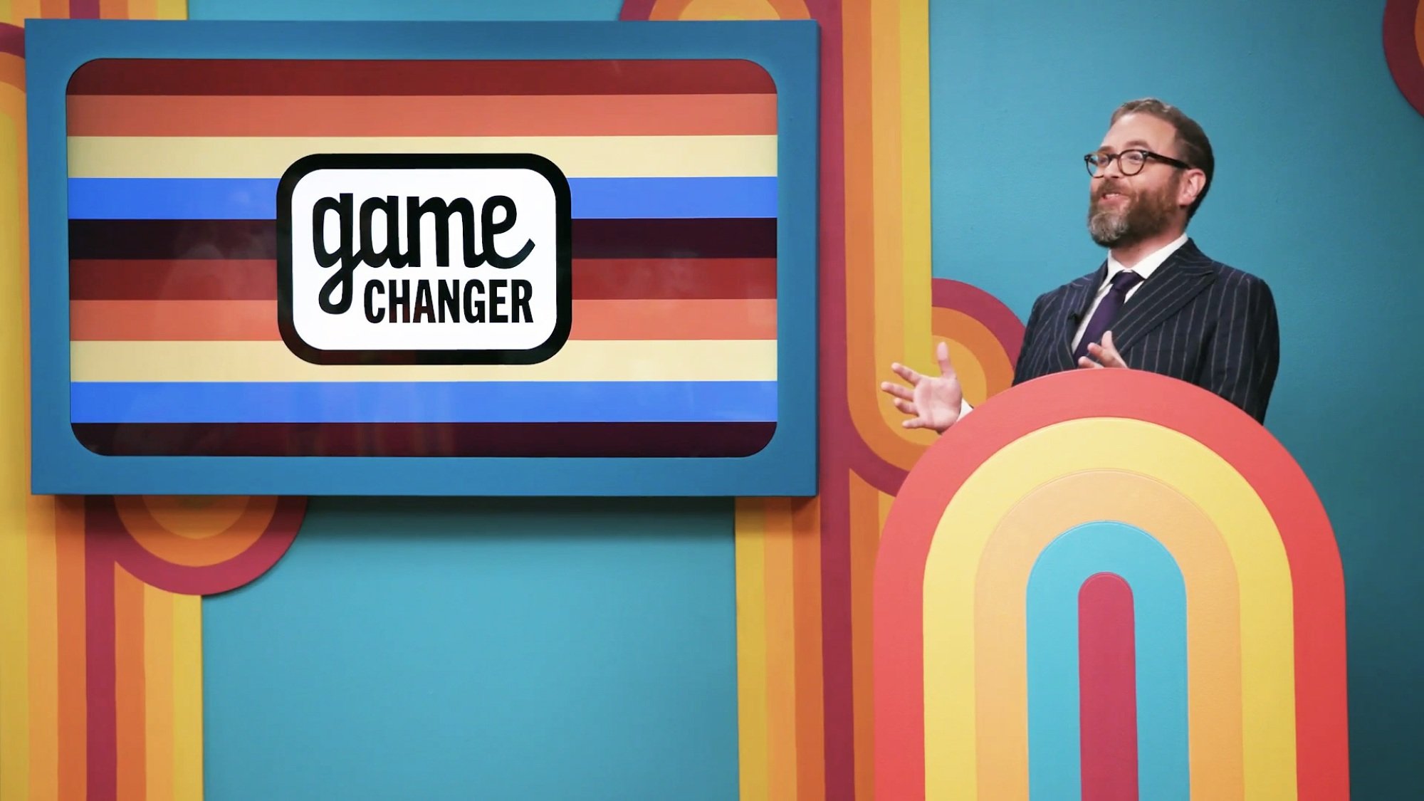 A game show host in a pinstripe suit stands next to a screen reading "Game Changer."