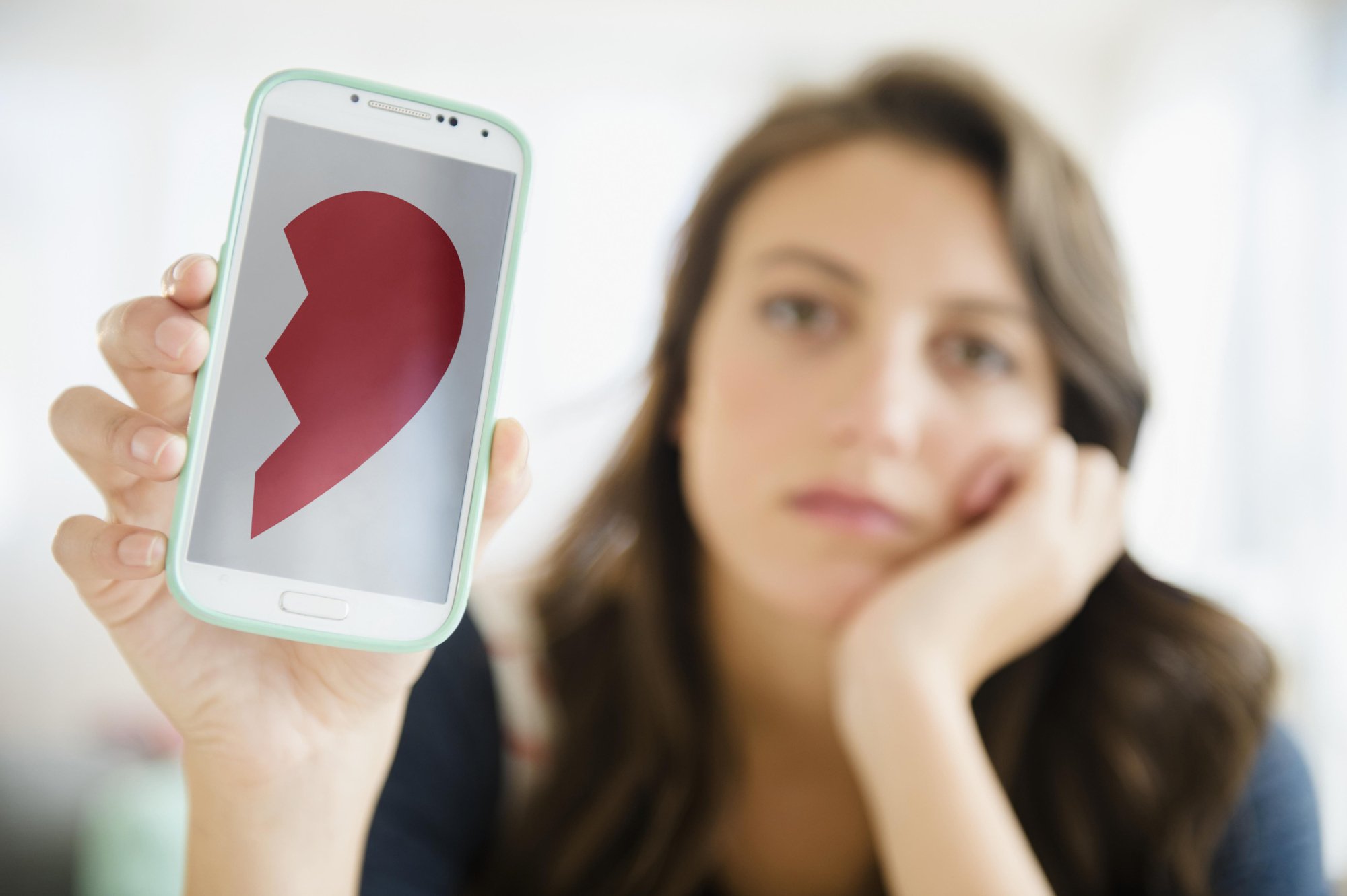 A sad looking woman holds up her phone with half a broken heart on the screen. 