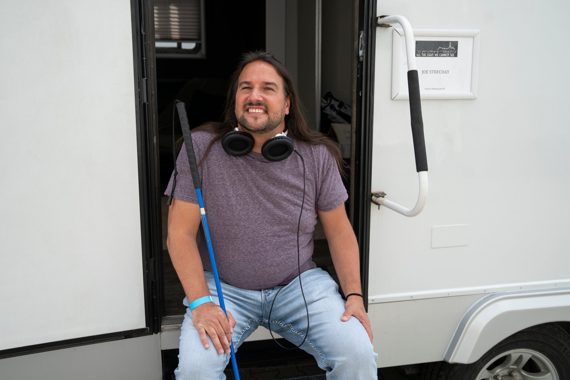 Strechay sits on the steps of a white set trailer. He is smiling at the camera, wearing a pair of large headphones around his neck and holding a blue cane.