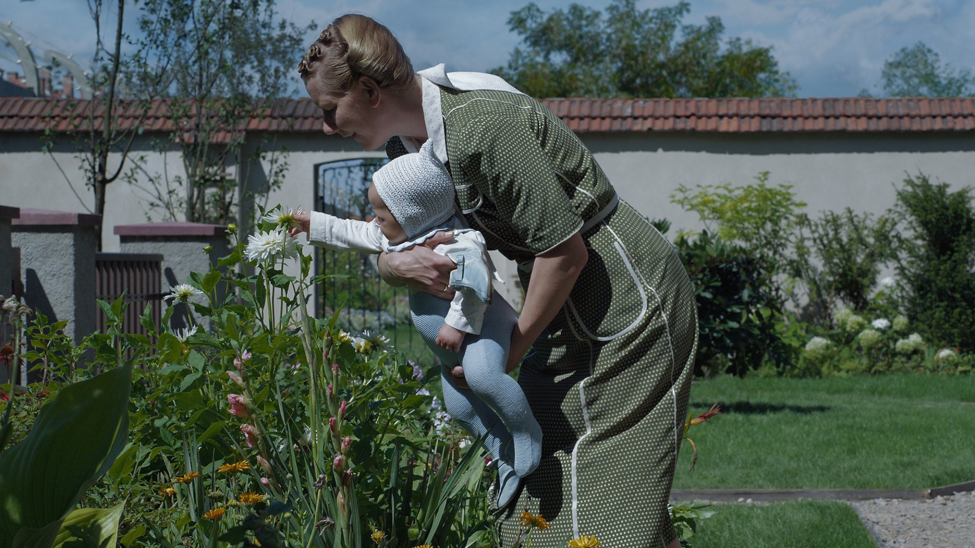 A woman holds a baby toward a flower in a pleasant walled garden.