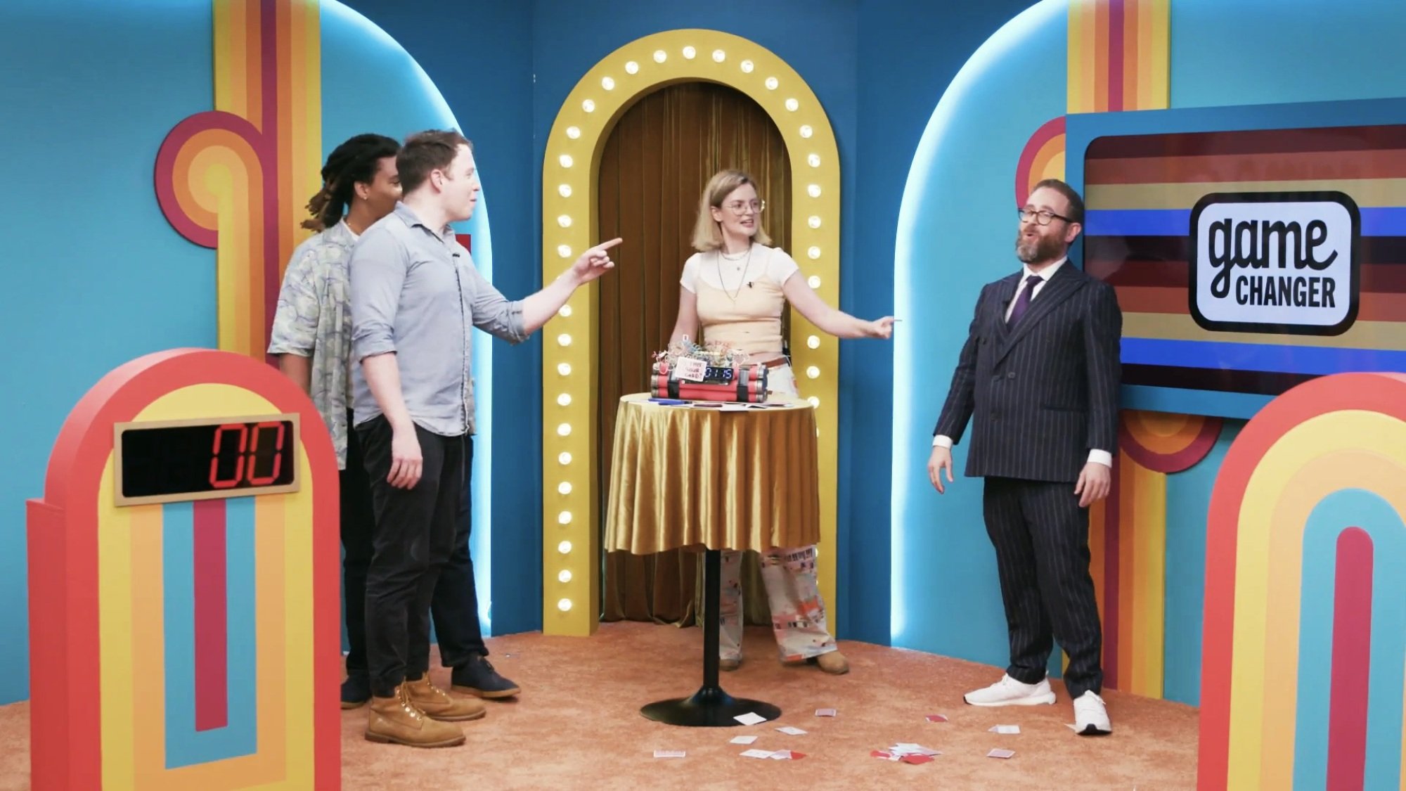 A group of game show contestants berate the host while standing around a fake pipe bomb.