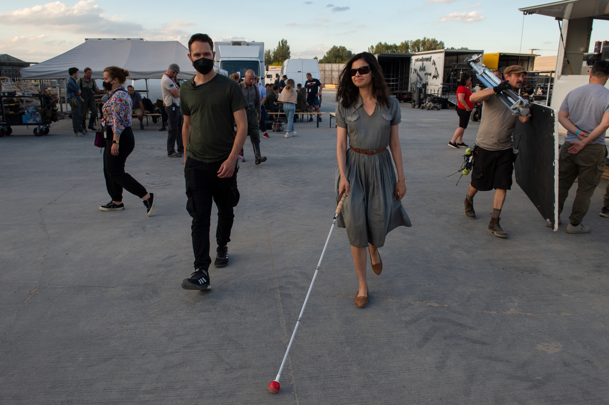 Loberti walks across a busy production set, wearing dark sunglasses and using a white cane.