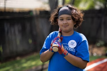 A child holding a baseball bat, wearing a blue Make-A-Wish shirt.