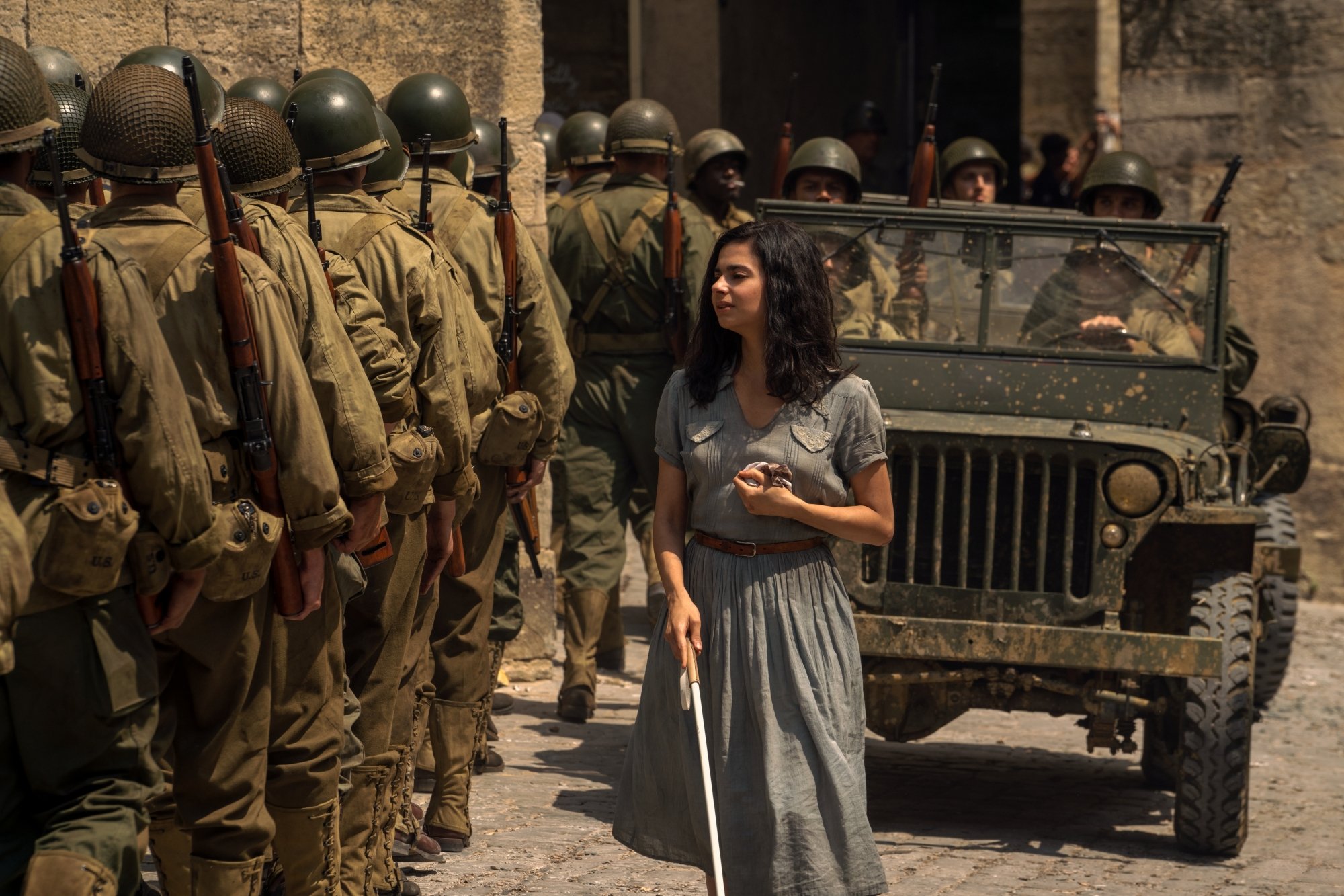 Loberti's character Marie-Laure walks by a line of soldiers and a military jeep. She appears in distress. 