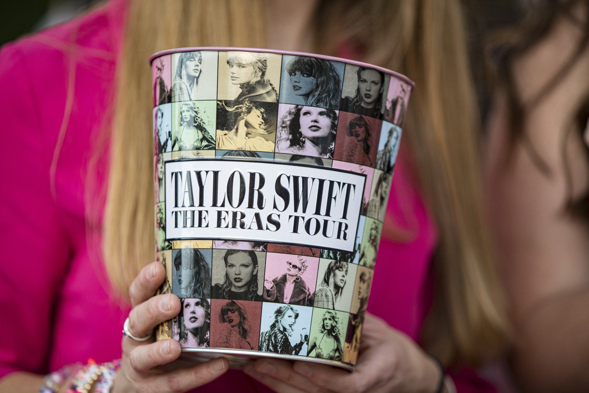 A white woman with blonde hair holds a popcorn bucket that reads, "Taylor Swift The Eras Tour."