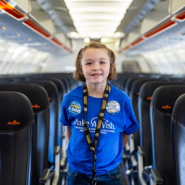 A child standing in the middle aisle of an airplane, wearing a blue Make-A-Wish shirt. 
