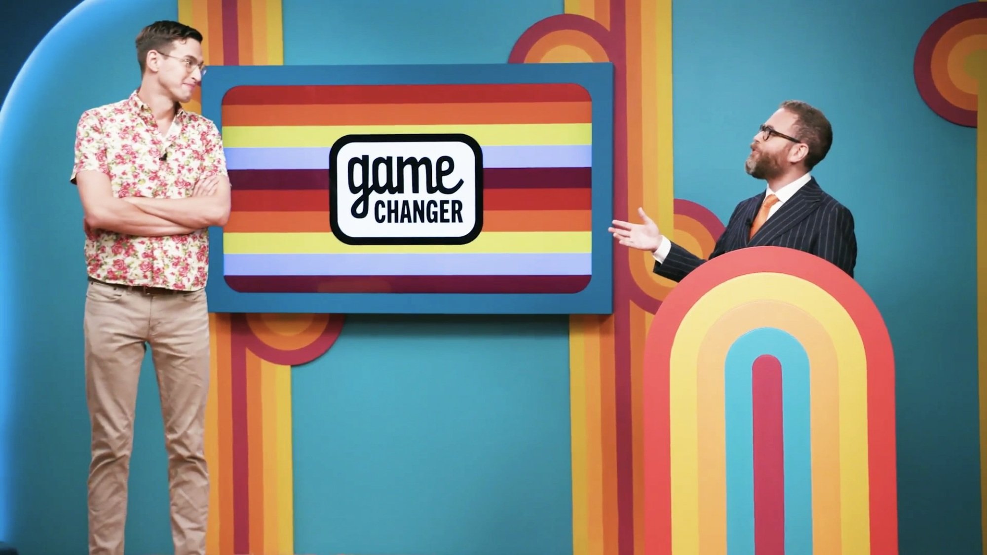 A game show contestant and host speak while standing by a screen that reads "Game Changer."