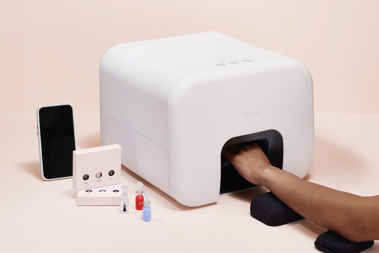 A hand in the Nimble manicure robot which is a white robot, next to a smartphone and nail polish capsules for the machine. 