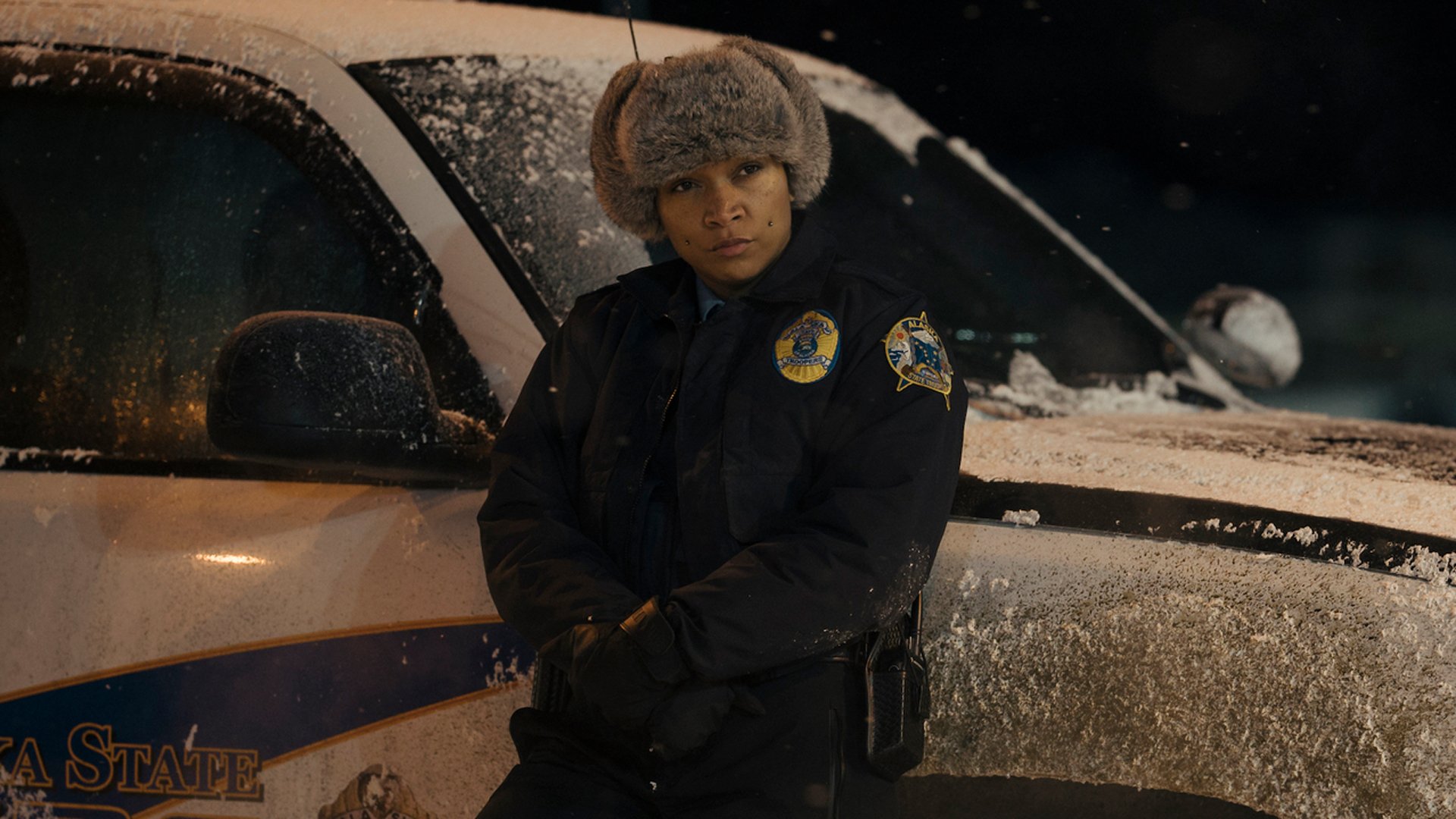 A detective leans on her car in the snow.