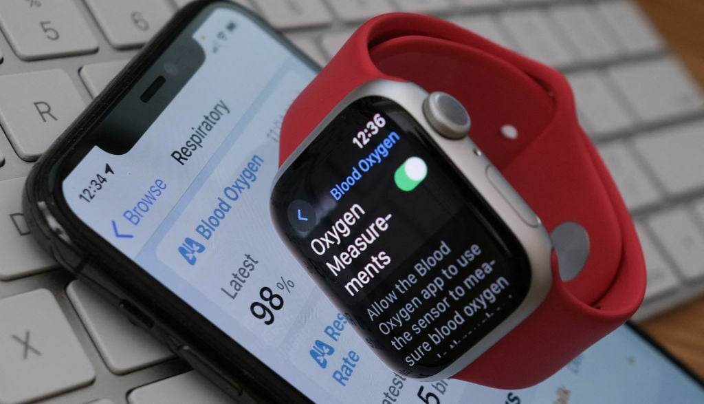 An Apple Watch 9 displaying the blood-oxygen level detection settings. It lies on top of an iPhone displaying respiratory data. Both lay on top of a keyboard.