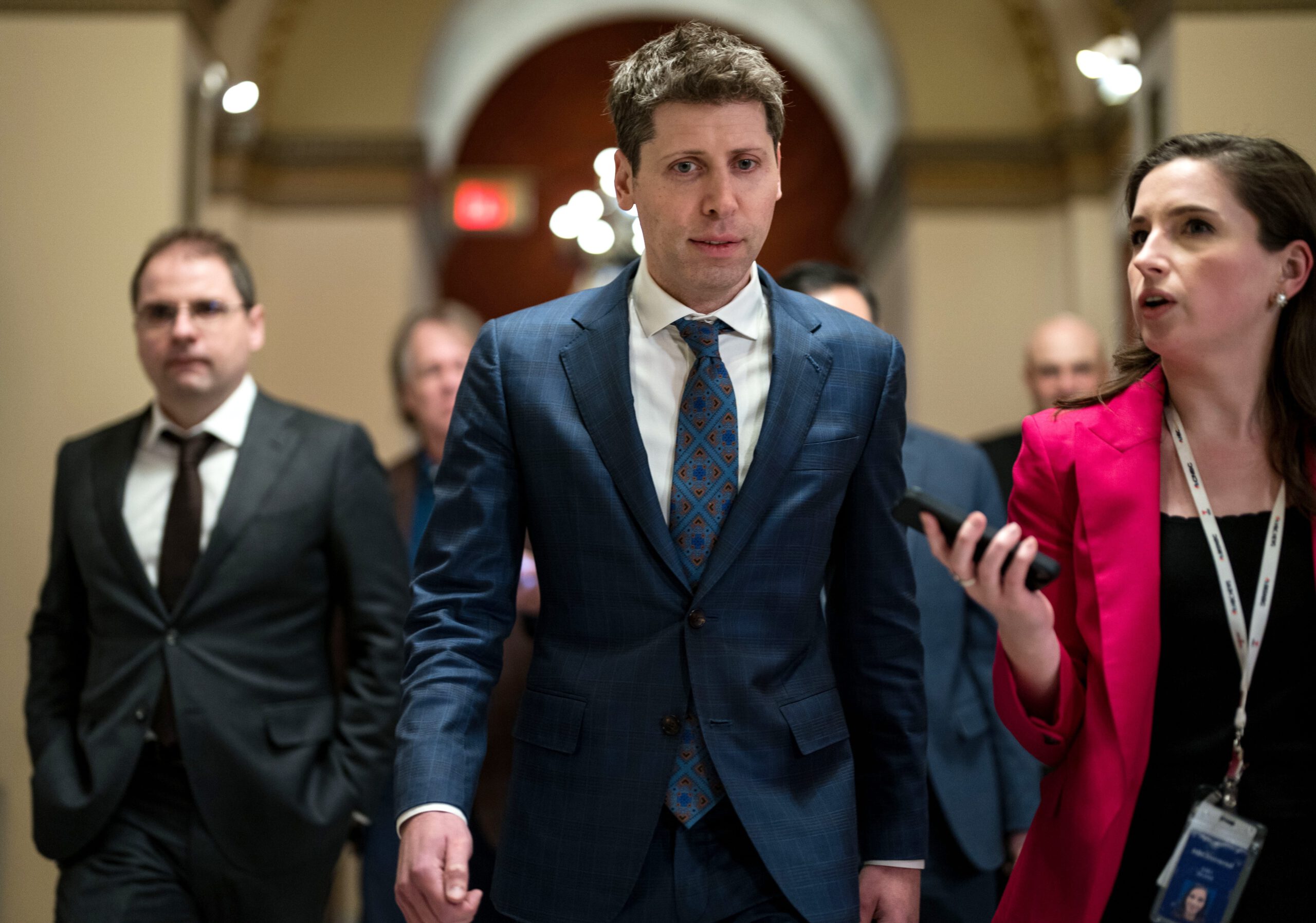 Sam Altman, center, in a dark blue suit, flanked to his left by a woman in red who appears to be speaking to him.