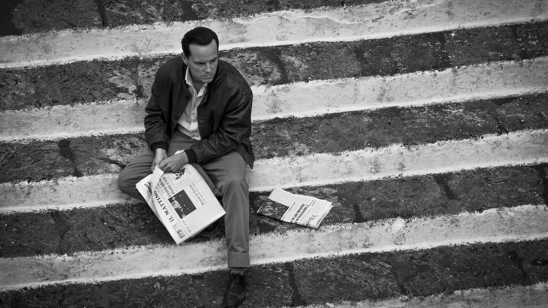A man sits on a public set of stairs with a newspaper in a black and white film still.