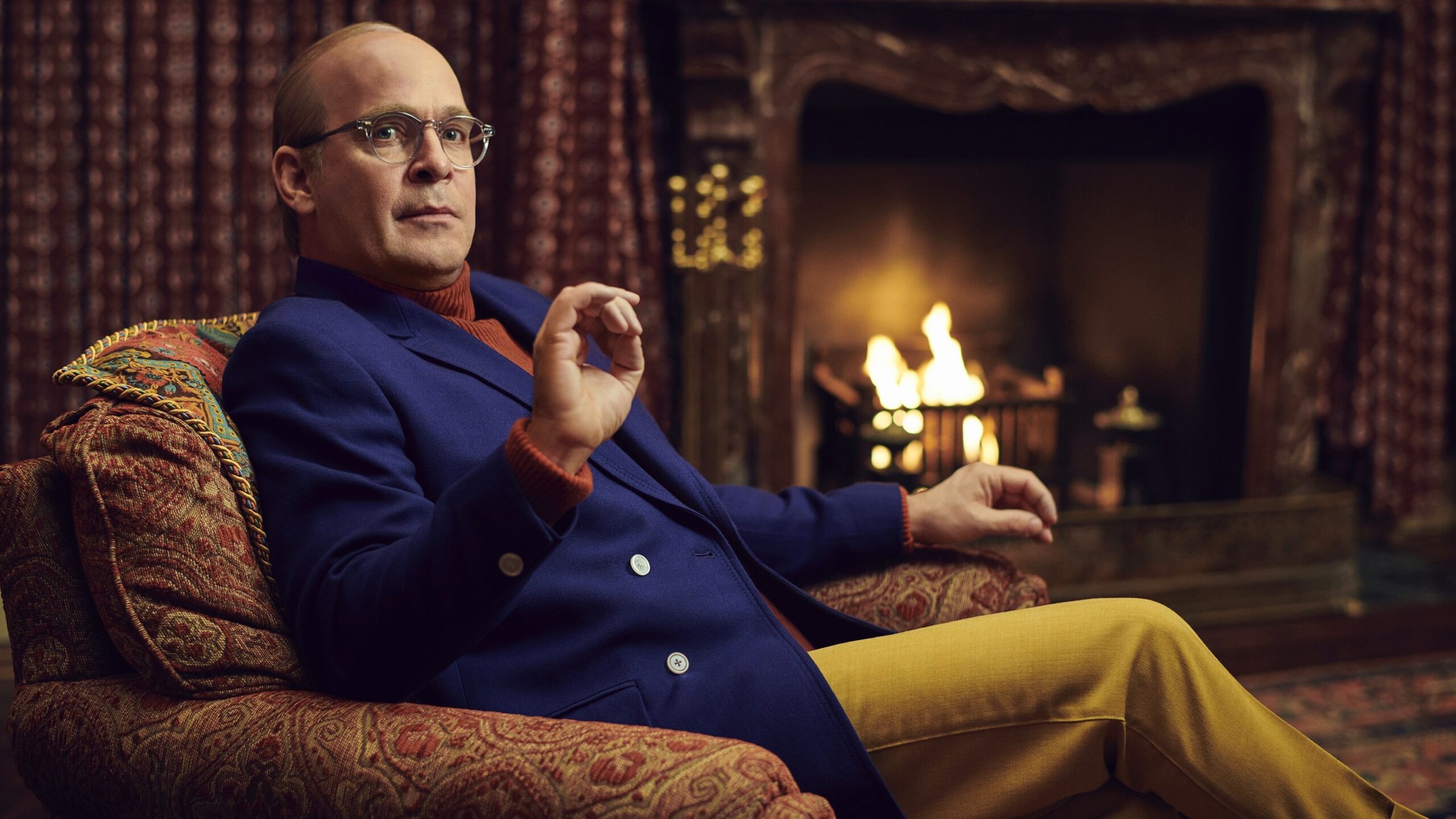 A man in a blue suit jacket, red turtleneck, and yellow pants sits in an armchair by a fireplace.