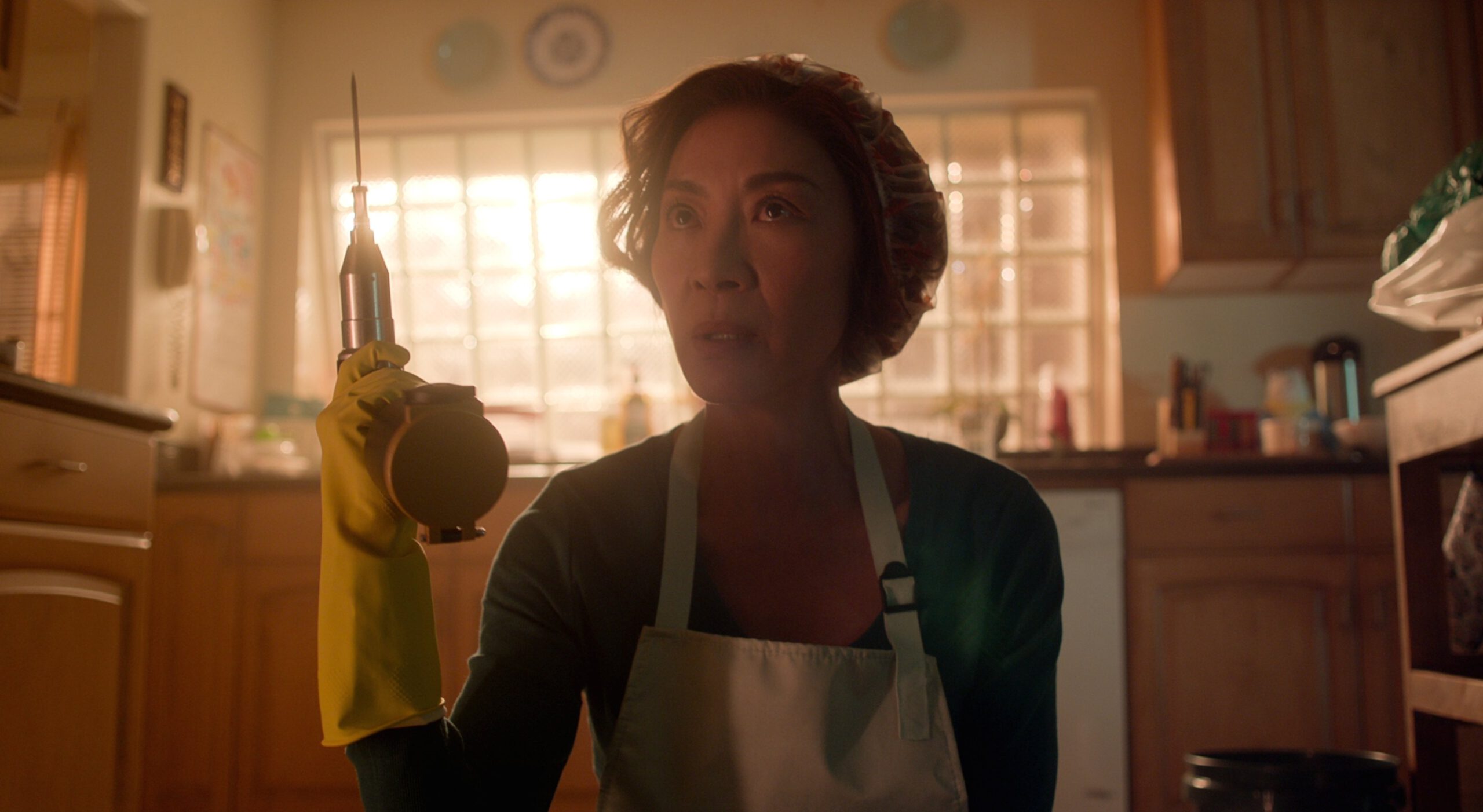 A woman in a hairnet and apron holds up a drill while kneeling on the kitchen floor.