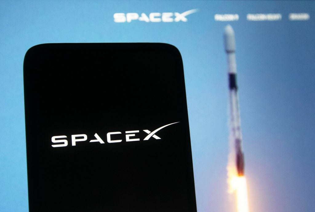 A SpaceX logo seen displayed on a smartphone. Behind it is an image of a rocket launching.