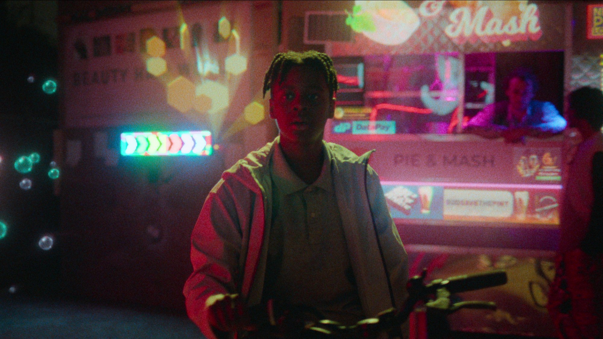 A teen boy stands with his bike in a neon-lit street.