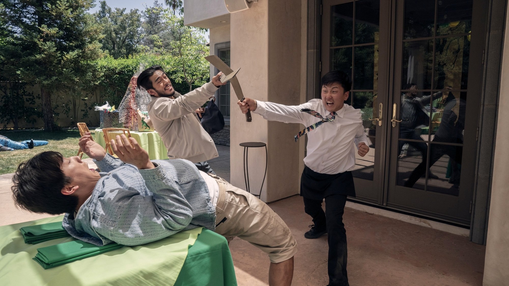 Two men fight with knives over a cowering man lying on a patio table.