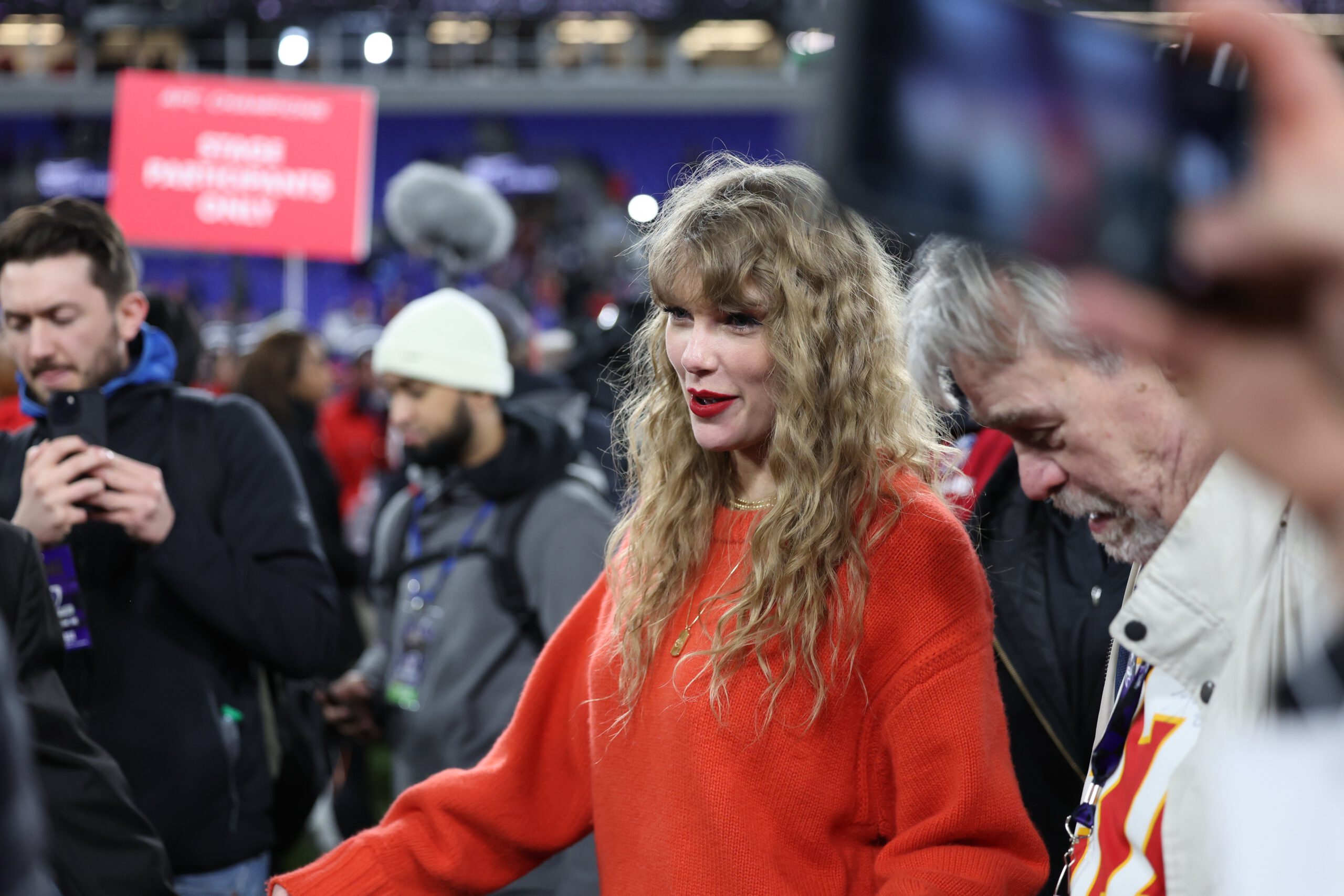Taylor Swift walking across the football field in a red sweater. 