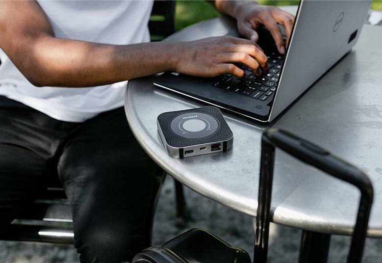a netgear nighthawk mobile hotspot sits on a table with someone working next to it on a laptop and a suitcase near the table