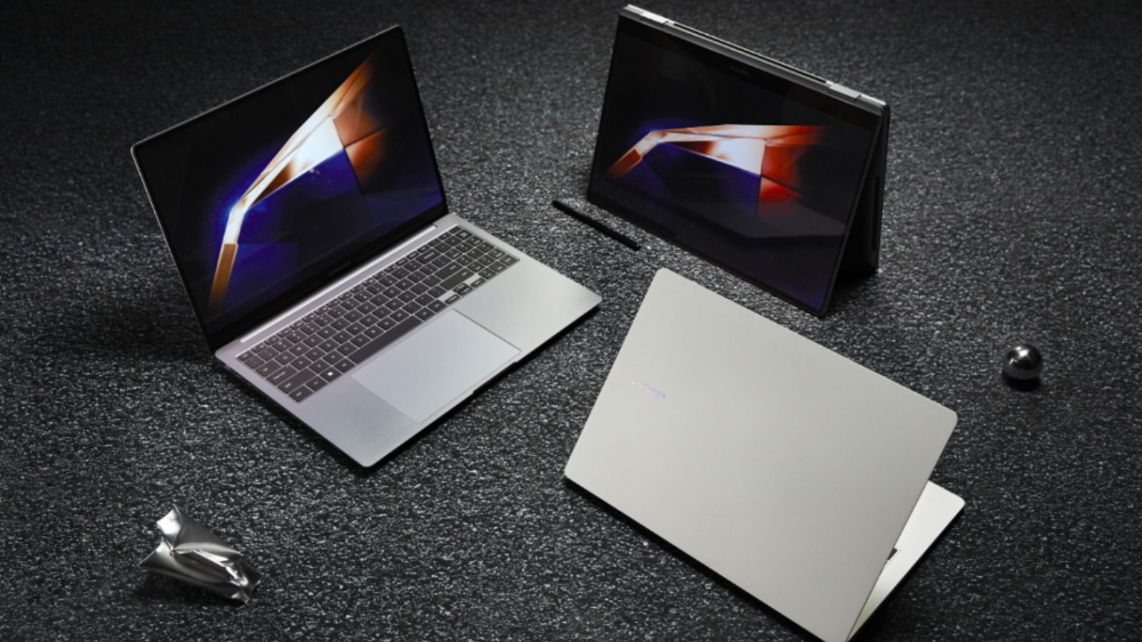 three laptops from the samsung galaxy book4 series on a gritty gray surface next to two pieces of metal