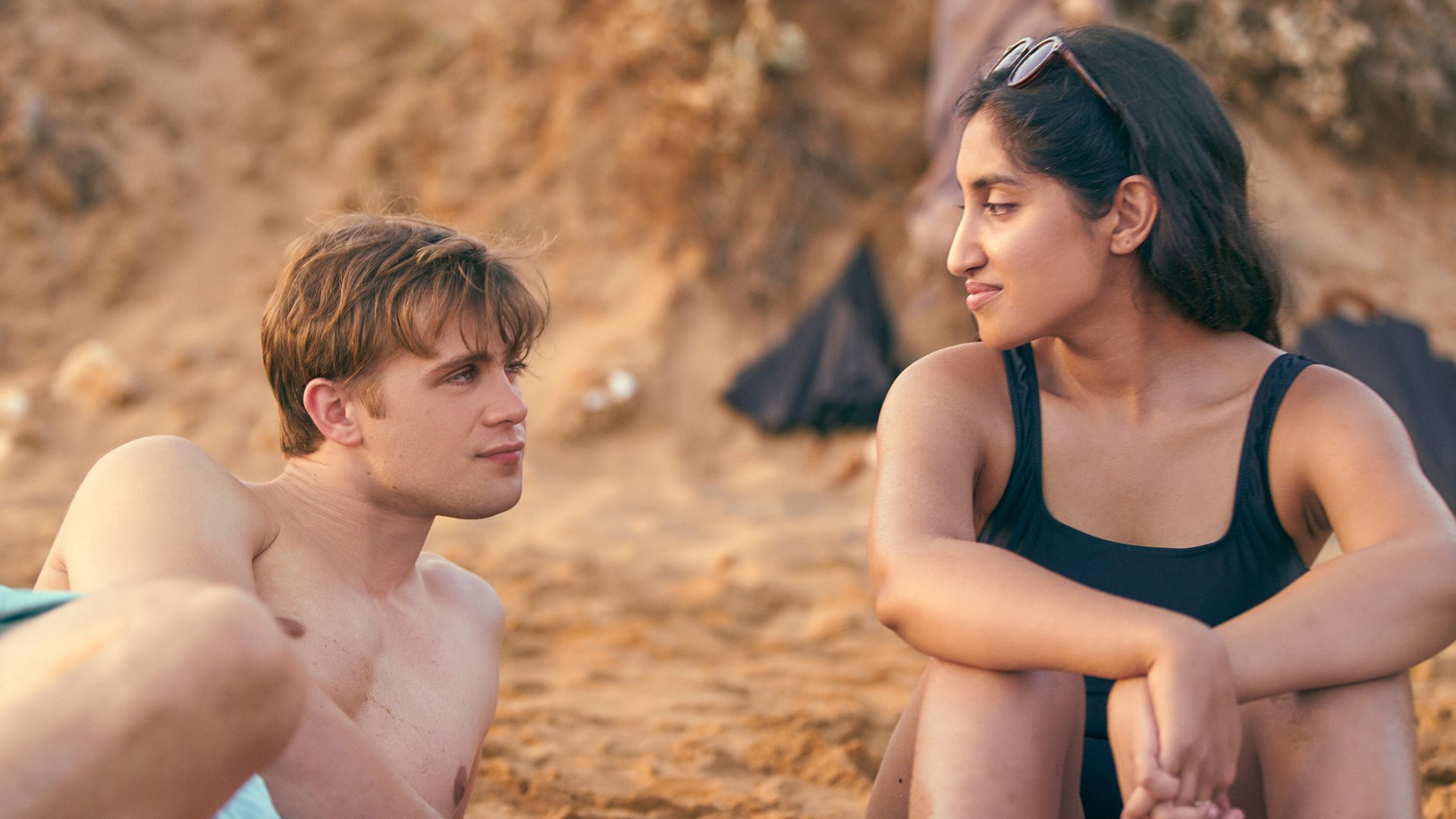 A young man and young woman sit on the beach looking romantically at each other.