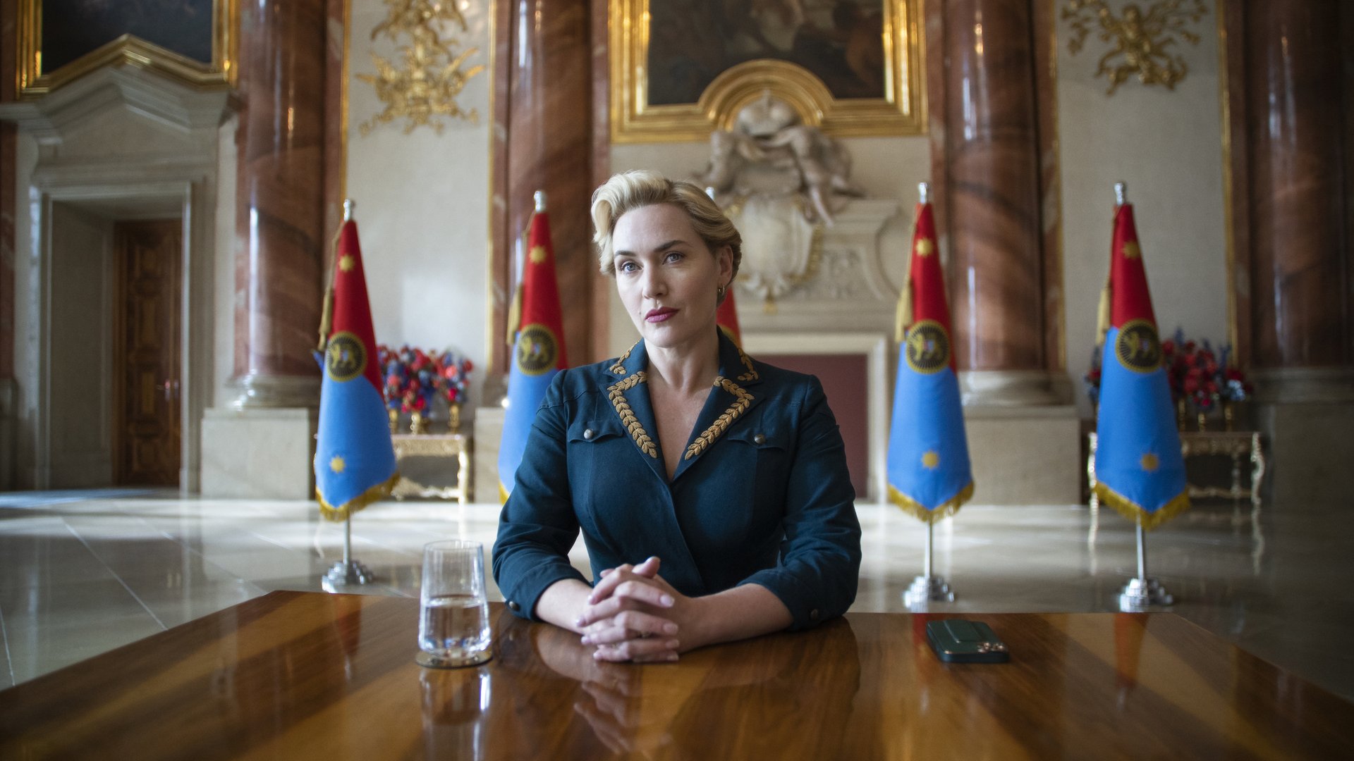 Kate Winslet sits at a desk dressed as a chancellor.