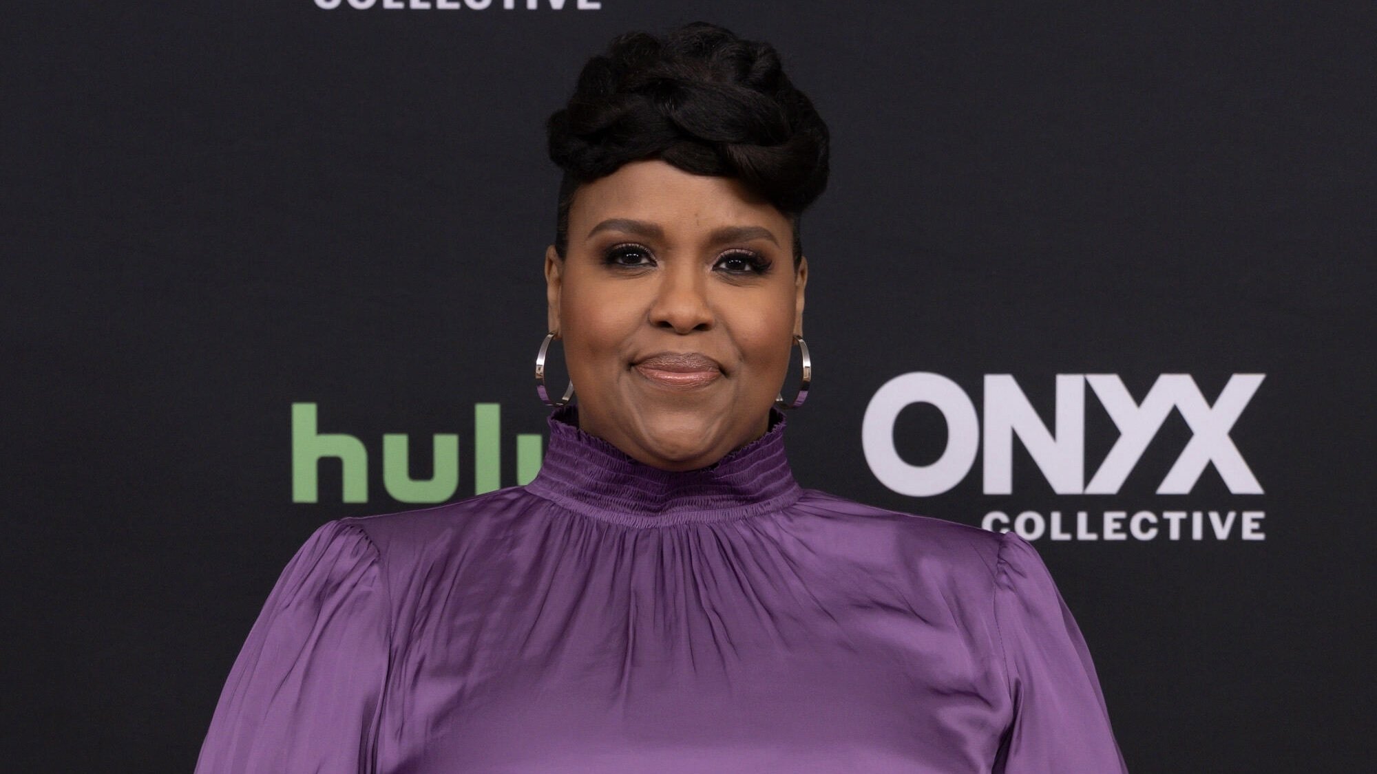 Natasha Rothwell wearing a purple dress and standing in front of a black backdrop that reads 