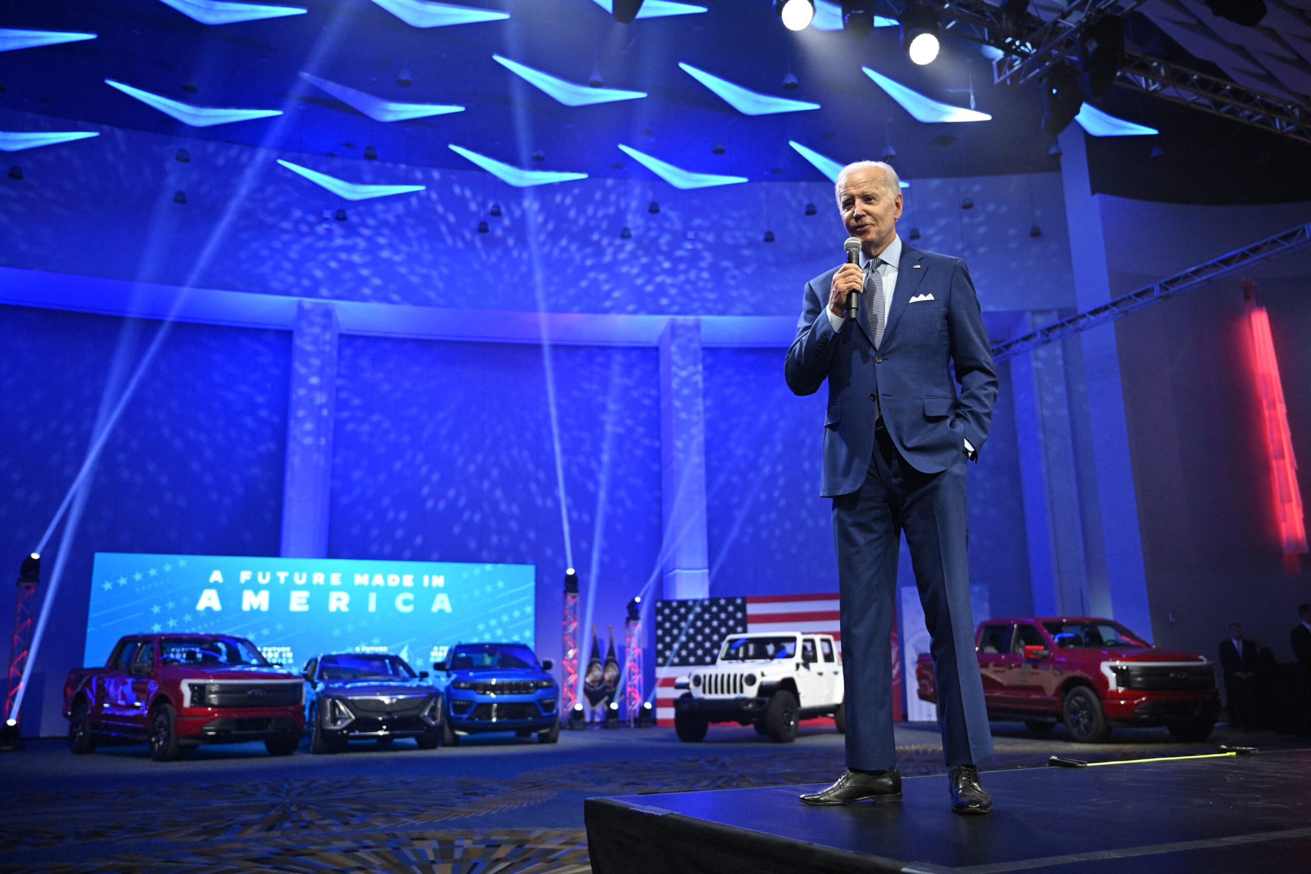 Old white man in blue suit speaks into a microphone. In the background is a display lined with electric vehicles in red, white, and blue variations