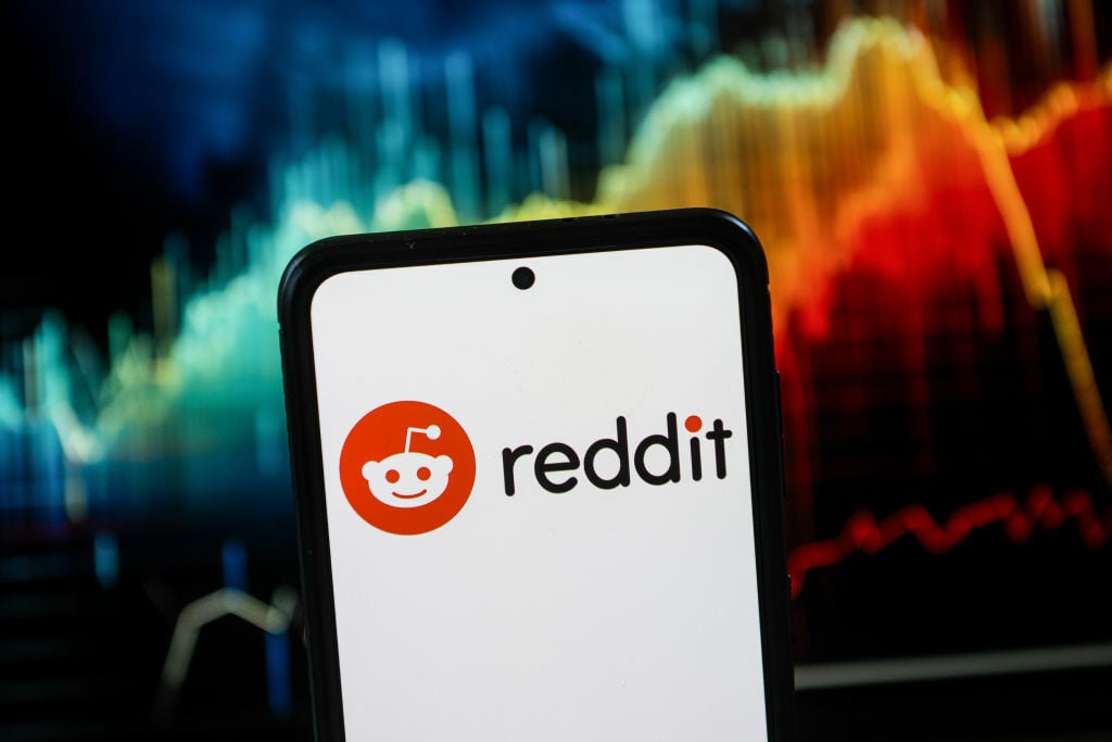 The Reddit logo on a smartphone, in front of a stock tracking graph.