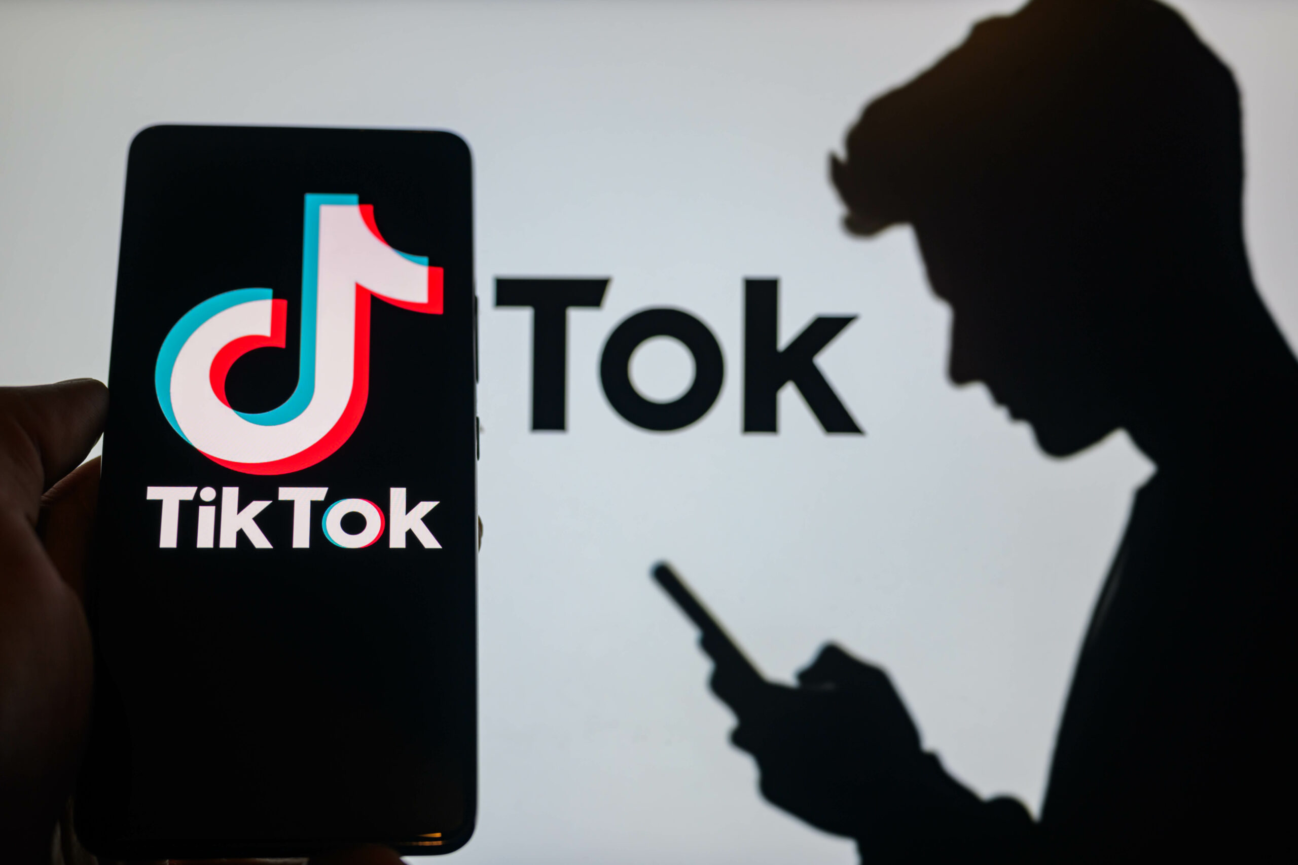 TikTok logo open on a phone screen next to the silhouette of a man looking at his phone. 