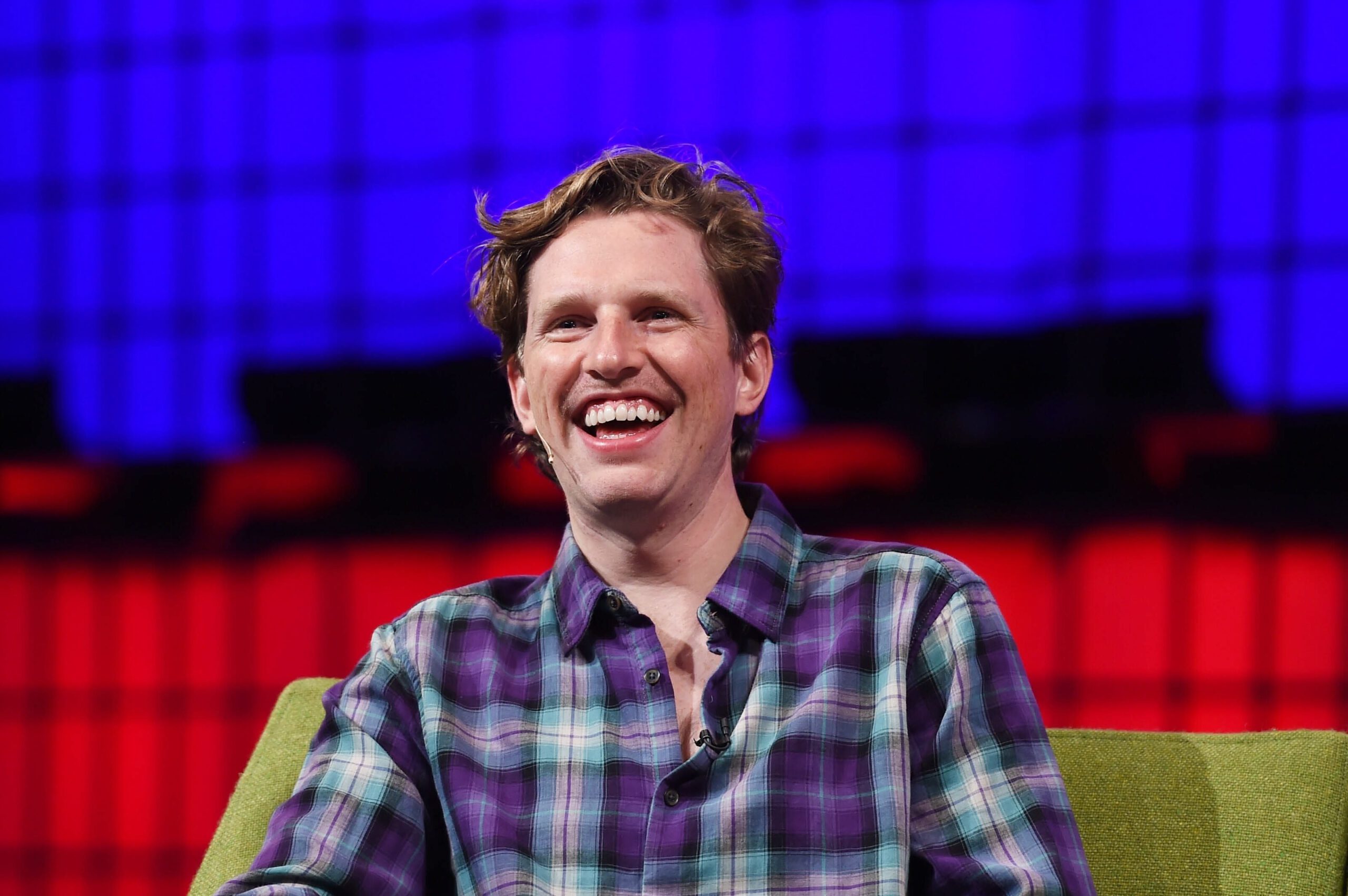 Matt Mullenweg is a white man with wavy blonde hair. He is wearing a plaid shirt and smiling on a lime green couch.