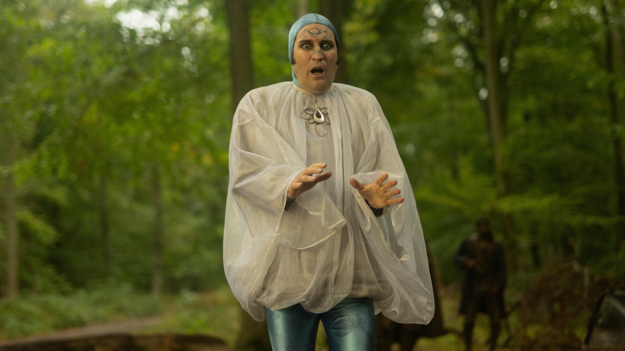 Noel Fielding dressed in a large white blouse and blue cap in a forest.