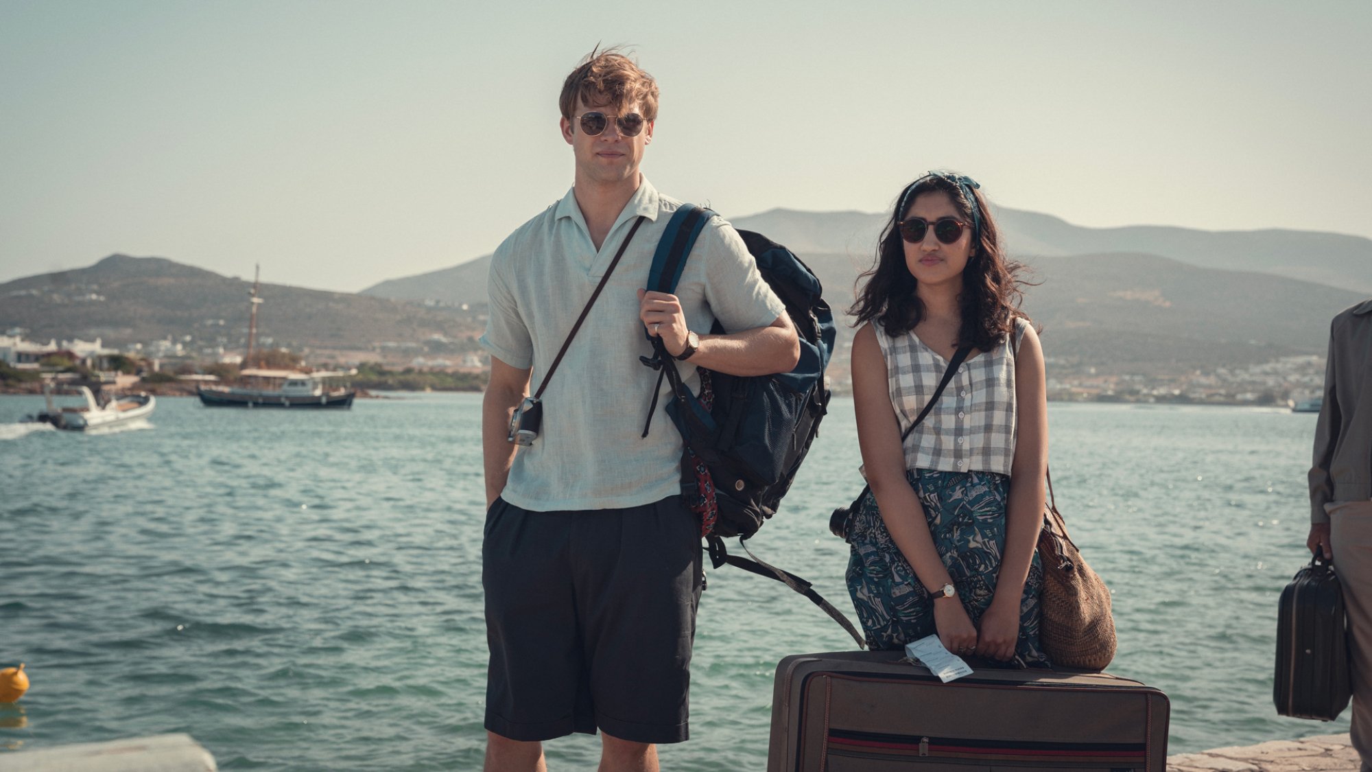 Two young people stand by the water in Greece holding their travel bags.