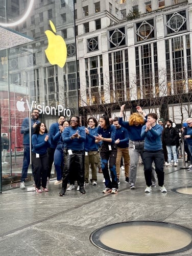 10 Apple employees in dark blue shirts cheer and clap in front of the Apple store.