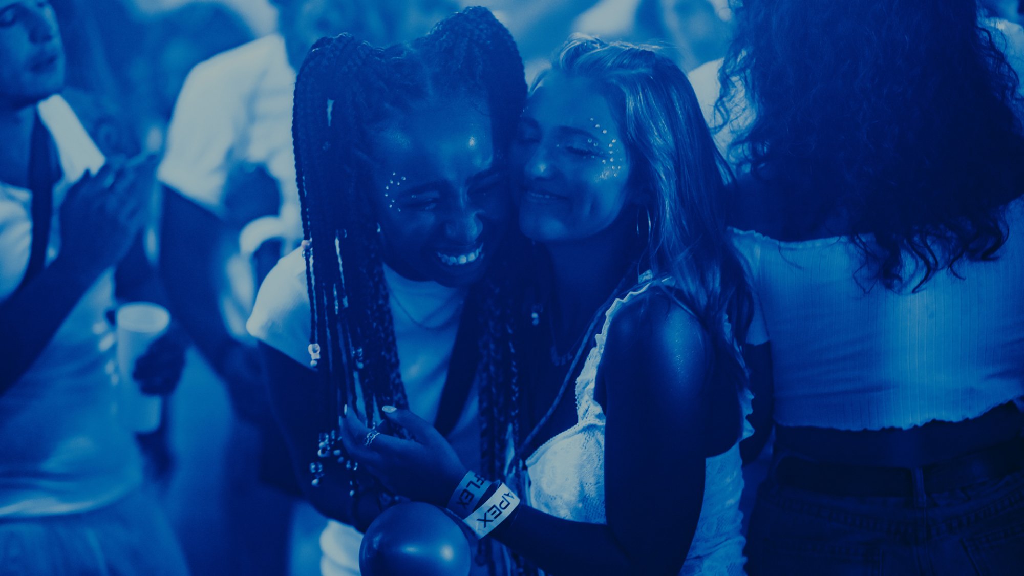 Two girls hug on a dancefloor with their eyes closed smiling.