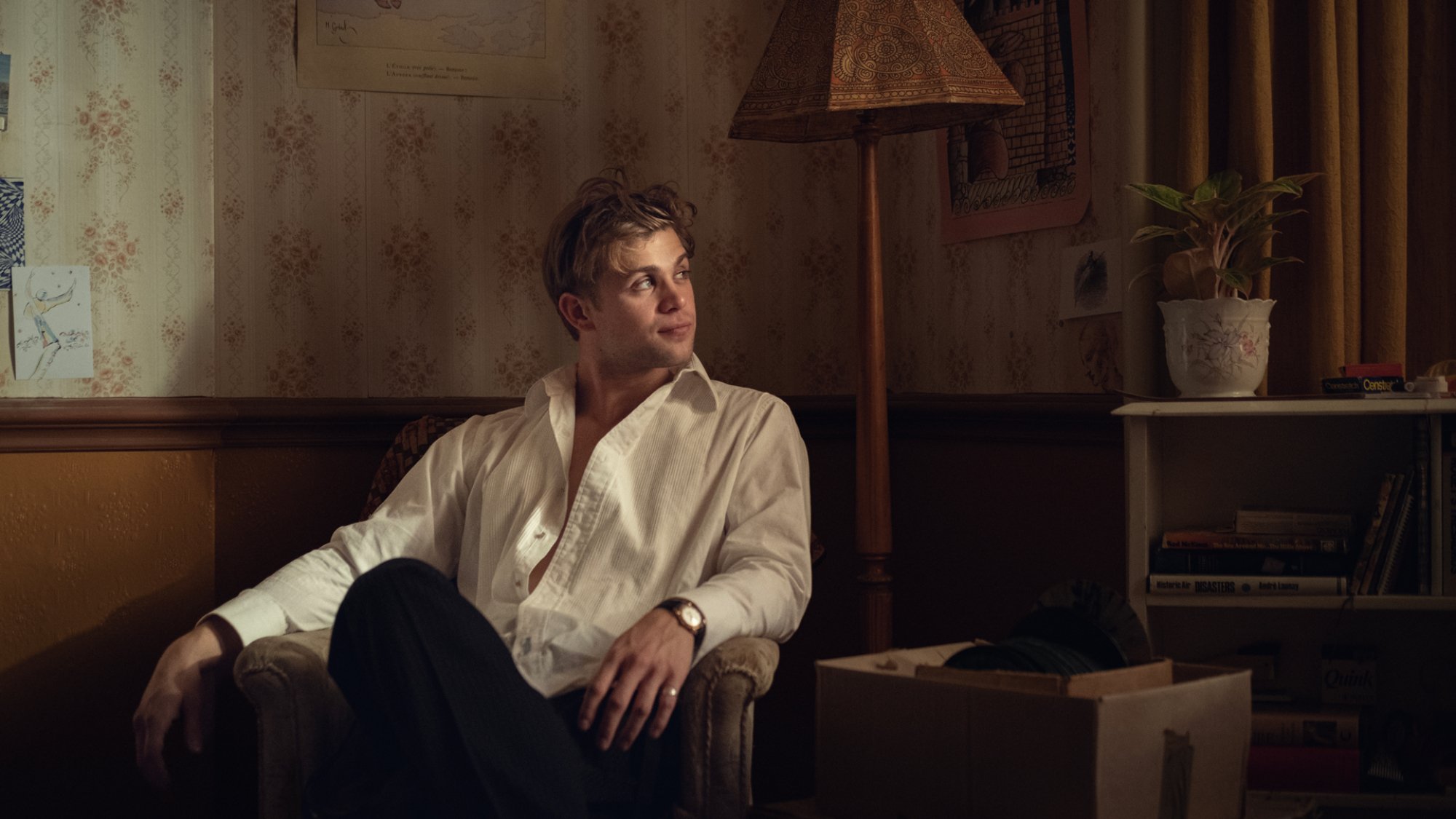 A young man in a white shirt sits in a dimly lit but opulent room.