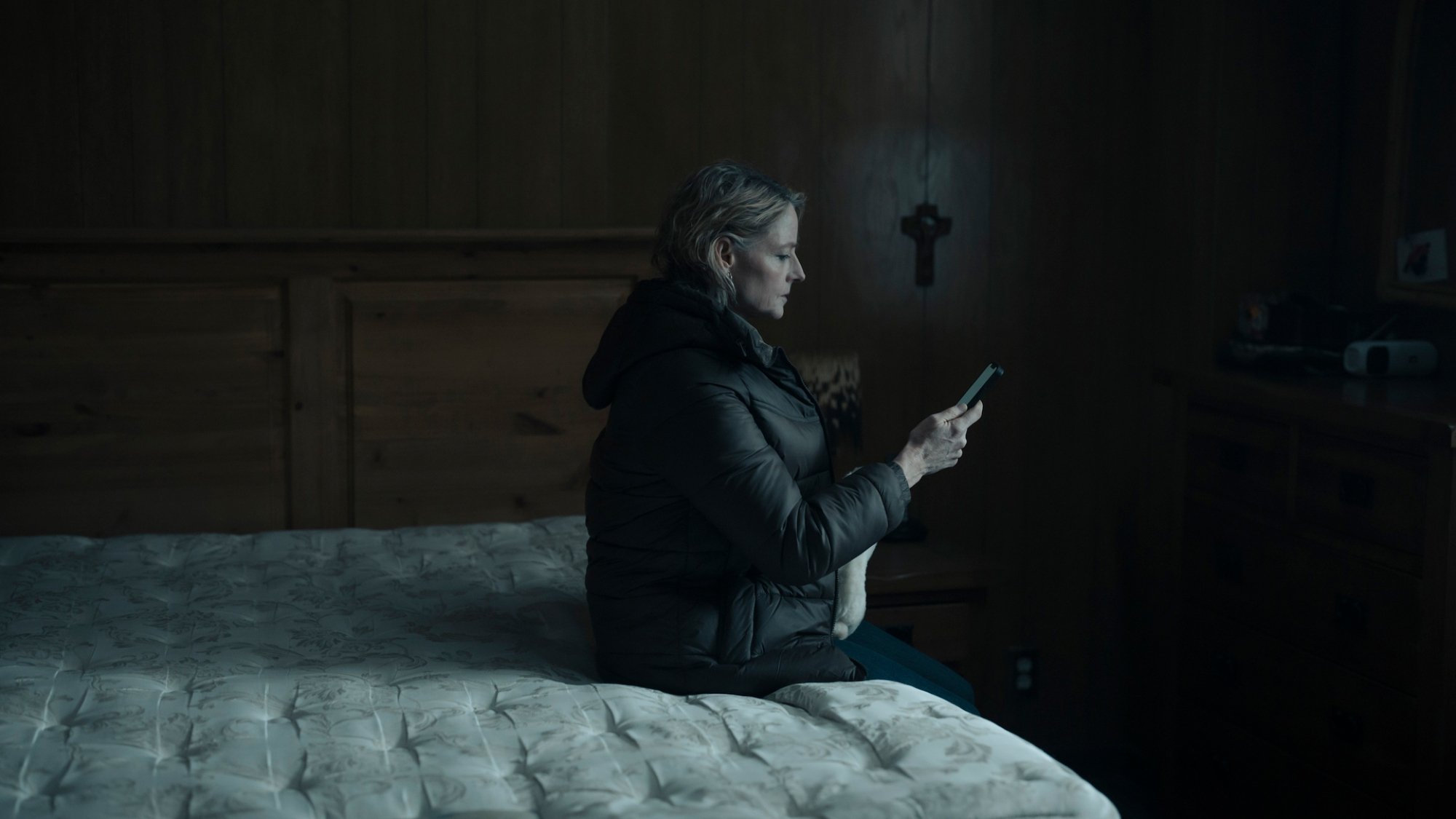 Jodie Foster sits on a stripped bed holding a phone.