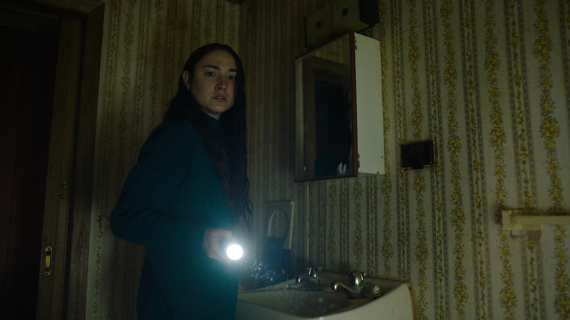 A woman in a dingy bathroom shines a torch off-camera, while looking afraid.