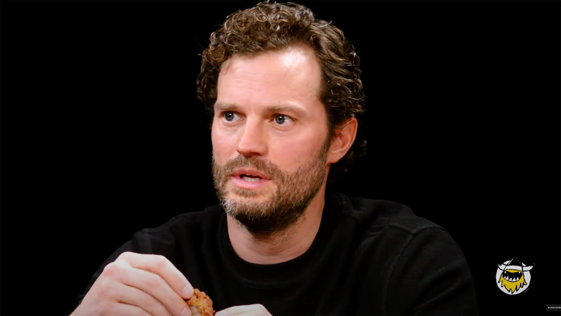 A man in a dark room holds a chicken wing while looking uncertain.