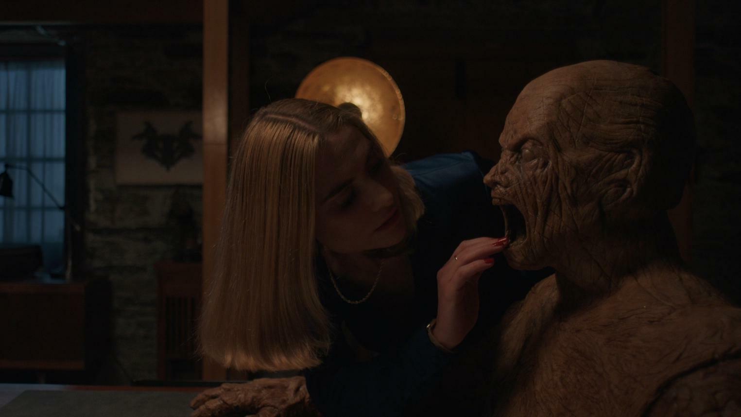 A woman reaches her hand into the mouth of a wooden man. 