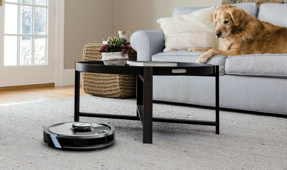 a shark matrix plus robot vacuum cleans the carpet in a living room that also has a table and a dog laying on a couch